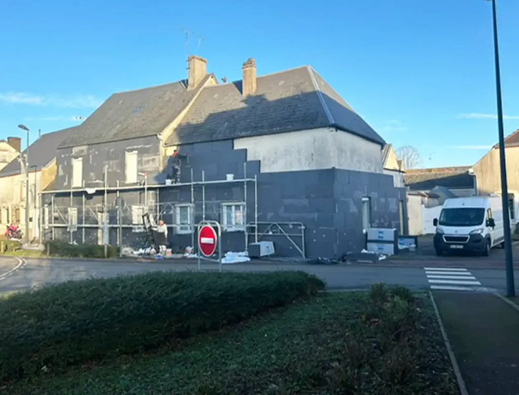 Une maison en cours de r&eacute;novation sur un chantier d'isolation par l'ext&eacute;rieur, avec des &eacute;chafaudages et des panneaux d'isolation sur les murs ext&eacute;rieurs. Des mat&eacute;riaux de construction sont &eacute;parpill&eacute;s sur le sol, une camionnette blanche est gar&eacute;e &agrave; proximit&eacute; et un panneau d'interdiction d'acc&egrave;s se trouve devant sous un ciel d&eacute;gag&eacute;.