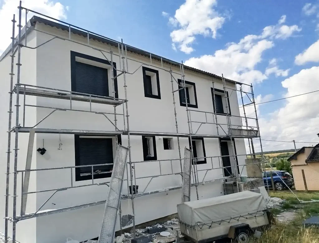Un bâtiment blanc de deux étages aux volets noirs est entouré d'un échafaudage métallique, typique d'un chantier de ravalement. Des matériaux de construction et une remorque couverte se trouvent au premier plan, sous un ciel partiellement nuageux.