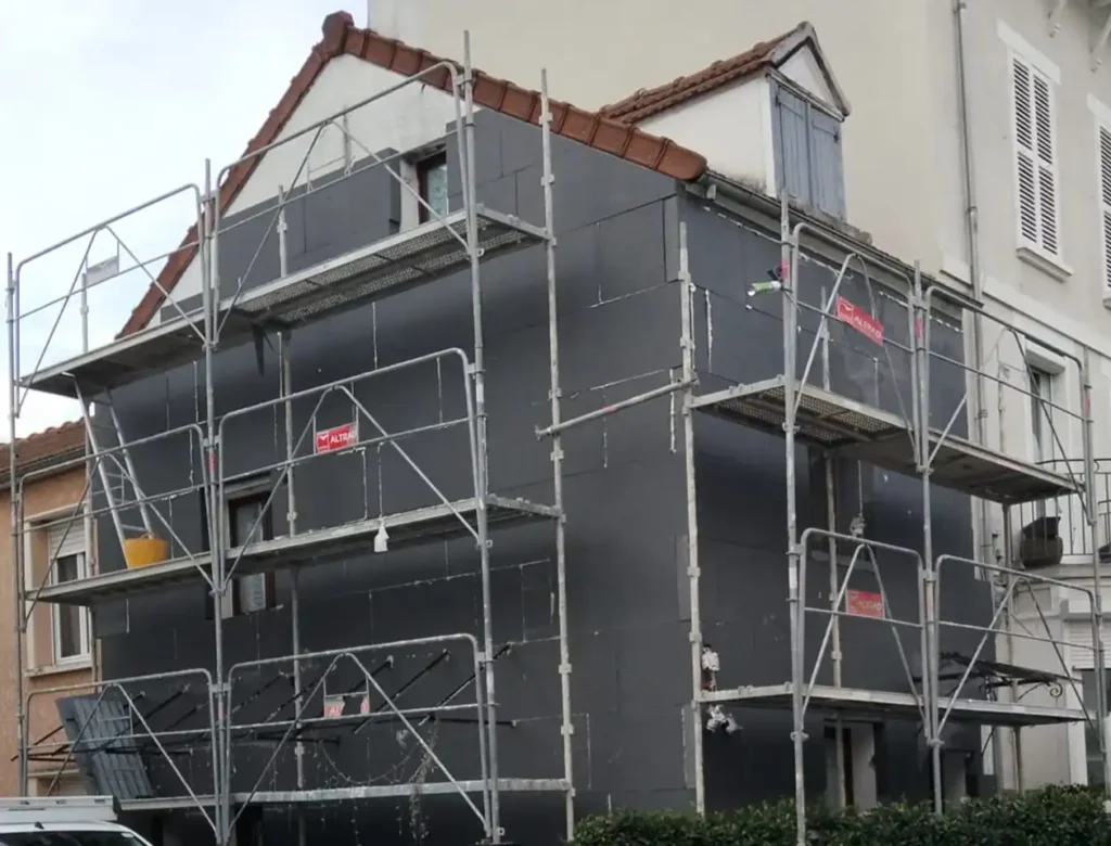 Une maison de deux &eacute;tages avec un toit en pente est recouverte d'un &eacute;chafaudage pour la construction ou la r&eacute;novation. Le b&acirc;timent, qui fait partie d'un chantier d'isolation thermique (ITE), est recouvert de panneaux isolants.