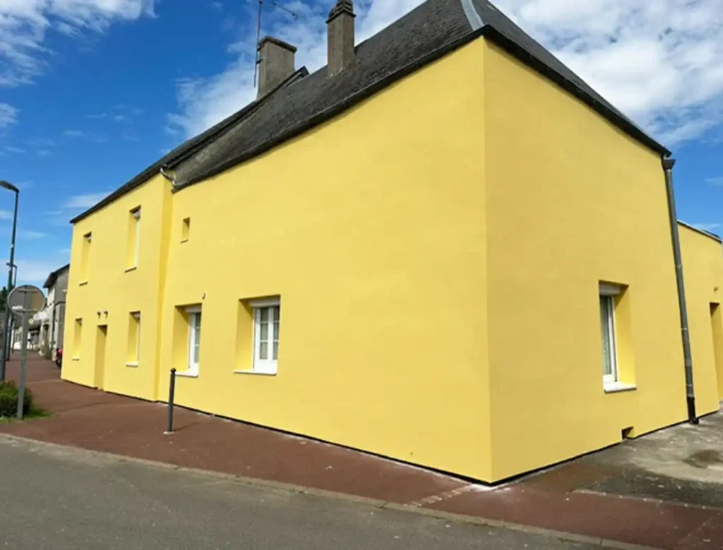 Une maison jaune vif avec des fen&ecirc;tres &agrave; cadre blanc se trouve au coin d'une rue sous un ciel partiellement nuageux. Avec une fin de chantier isol&eacute;e par l'ext&eacute;rieur, le b&acirc;timent simple et moderne pr&eacute;sente des murs lisses et un toit gris fonc&eacute;.