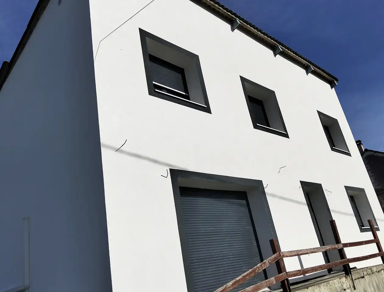 Ravalement de façade maison rurale – Isolex Bâtiment moderne blanc avec trois fenêtres rectangulaires et un grand volet roulant gris au rez-de-chaussée. Une clôture en bois longe la façade, mettant en valeur le ravalement de façade frais sous un ciel bleu clair.