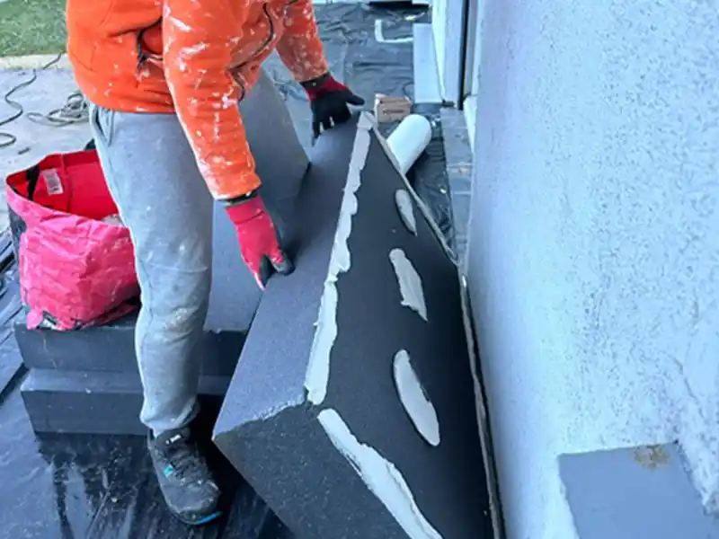 Application panneau isolant - Isolex Une personne portant une veste orange et un pantalon gris manipule l'application de panneau isolant, plaçant un panneau isolant avec de l'adhésif sur un mur extérieur. Un sac rouge et d'autres matériaux de construction se trouvent à proximité.