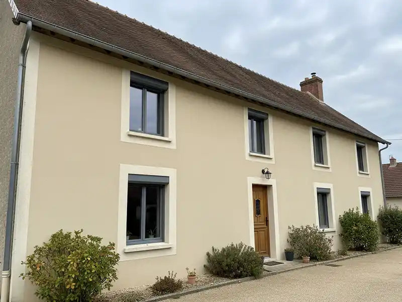 Isolation extérieure maison rurale - Isolex Une maison beige à deux étages avec un toit en pente marron, comportant une isolation extérieure, quatre fenêtres, une porte centrale en bois et de petits buissons le long de la façade, sous un ciel nuageux.