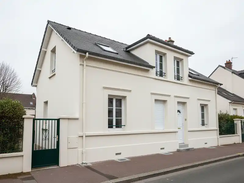 Maison &agrave; deux &eacute;tages de couleur cr&egrave;me, fra&icirc;chement repeinte, avec un toit gris fonc&eacute; et plusieurs fen&ecirc;tres &agrave; cadre blanc, situ&eacute;e dans une rue calme avec un portail vert et un trottoir devant.